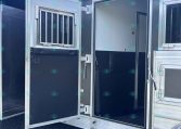 Open aluminum trailer door revealing the dark interior and a small barred panel on the left; trailer sits on gravel with blue sky above, zipper-like step at the bottom edge.
