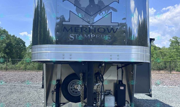Metal horse trailer with Merhow Stampede logo in front, parked on gravel under a blue sky