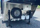 Open trailer storage area with spare tire, metal toolbox, two propane tanks, and stabilizing legs on gravel ground.