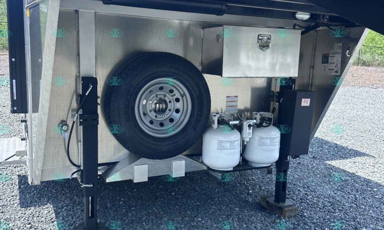 Open trailer storage area with spare tire, metal toolbox, two propane tanks, and stabilizing legs on gravel ground.