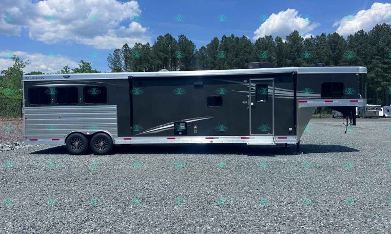 Side view of a dark gray Merhow horse trailer with two axles, parked on a gravel lot with trees in the background.
