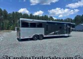 Large silver Merhow horse trailer with three axles and dark windows, parked on a gravel lot with pine trees and a blue sky in the background.