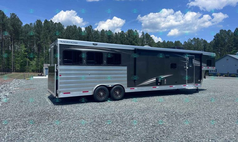 Large silver Merhow horse trailer with three axles and dark windows, parked on a gravel lot with pine trees and a blue sky in the background.