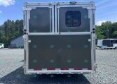 Rear view of a gray aluminum trailer with double doors, a small window, and metal latches, parked on a gravel lot.
