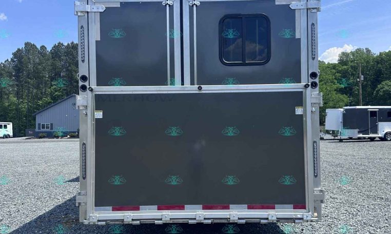 Rear view of a gray aluminum trailer with double doors, a small window, and metal latches, parked on a gravel lot.
