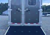 Rear view of a gray double-door trailer with a lowered loading ramp in a gravel lot, small window on the right door, blue sky above.