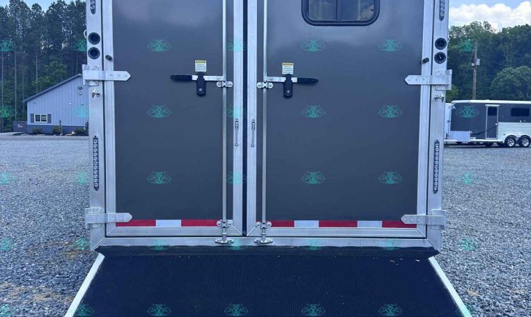 Rear view of a gray double-door trailer with a lowered loading ramp in a gravel lot, small window on the right door, blue sky above.