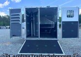 Open horse trailer with divider and stairs visible inside; ramp extended onto gravel lot, other trailers in background.