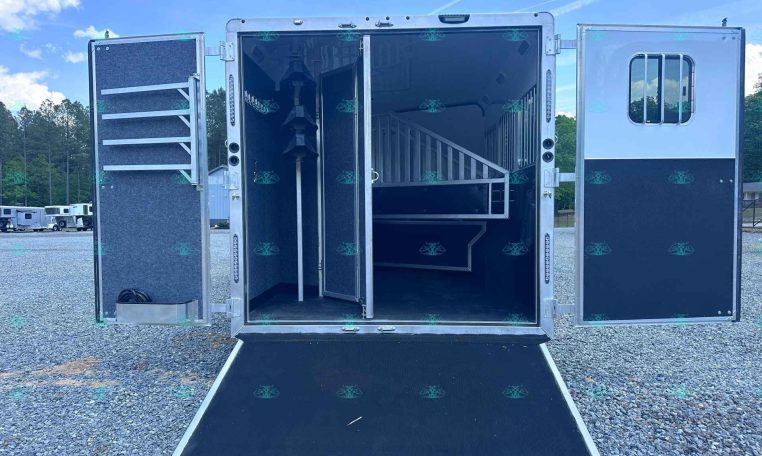 Open horse trailer with divider and stairs visible inside; ramp extended onto gravel lot, other trailers in background.