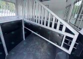Interior of a aluminum horse trailer with a partition and stair ramp, black padded walls, and windows along the side.