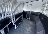 Interior of a horse trailer with dividing stalls, metal railing, and raised stairs along a white and black padded divider wall.