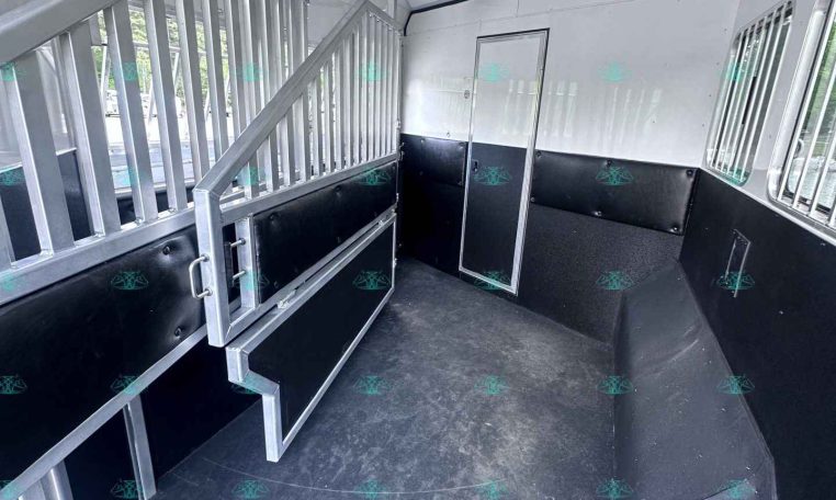 Interior of a horse trailer with dividing stalls, metal railing, and raised stairs along a white and black padded divider wall.