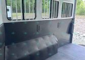 Interior of a horse trailer with barred windows along the side, gray walls, and a black rubber floor, showing a space for horses and ventilation.