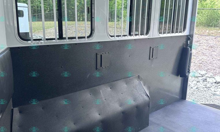 Interior of a horse trailer with barred windows along the side, gray walls, and a black rubber floor, showing a space for horses and ventilation.