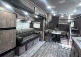 Interior of a modern trailer living area with gray upholstery, wooden cabinets, a sofa, and a small dining table with a vase of flowers.