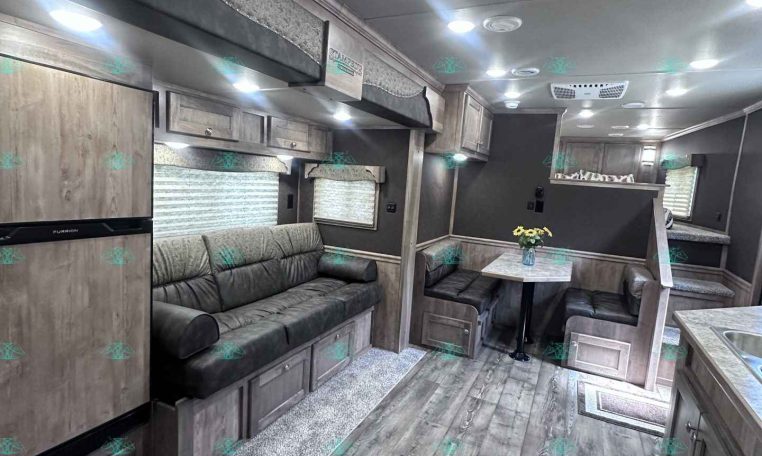 Interior of a modern trailer living area with gray upholstery, wooden cabinets, a sofa, and a small dining table with a vase of flowers.