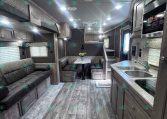 Interior of a modern trailer/RV showing a living area with a sofa, dining table, and kitchen along the right; wood-look flooring and gray cabinetry.