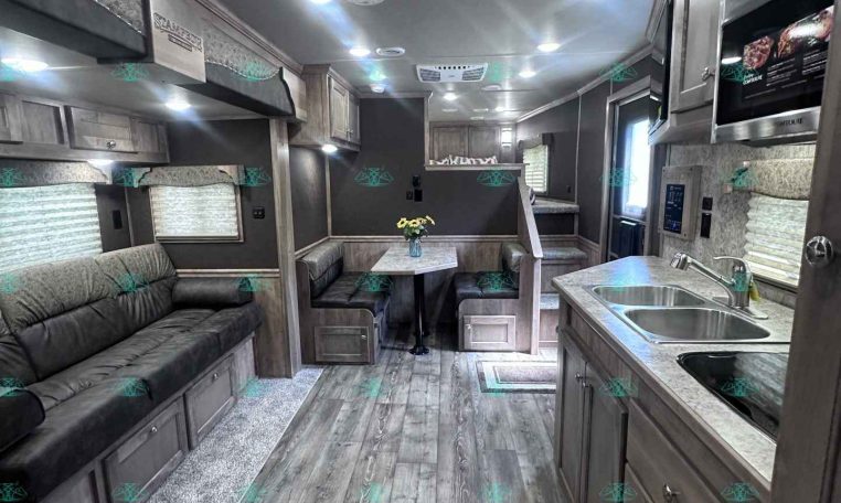 Interior of a modern trailer/RV showing a living area with a sofa, dining table, and kitchen along the right; wood-look flooring and gray cabinetry.