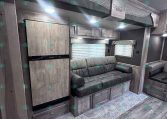 RV living area with built-in fridge, upper bunk, and gray wood cabinets along a windowed wall