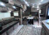 Interior of a modern grey-toned travel trailer living area with a long sofa, dinette, and overhead storage cabinets.