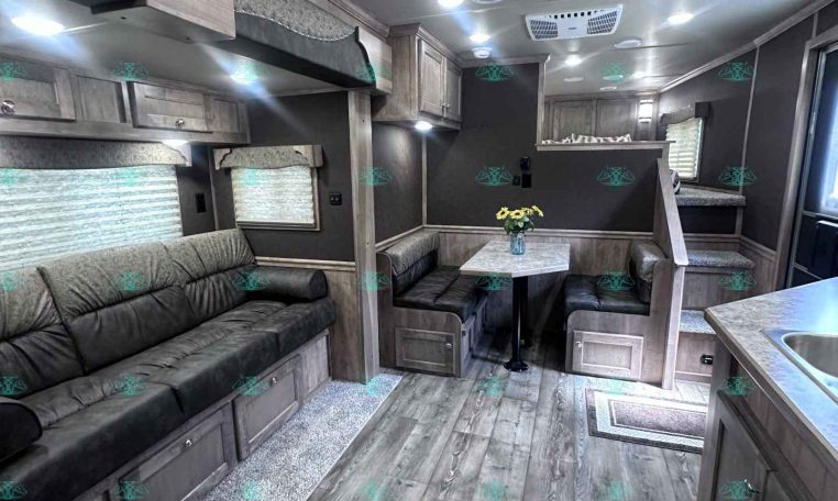 Interior of a modern grey-toned travel trailer living area with a long sofa, dinette, and overhead storage cabinets.