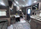 Interior of a modern RV trailer living area with dining booth, sofa, and overhead cabinets in gray wood tones.