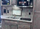 Kitchen area in a trailer: gray wood cabinets, stainless microwave, double sink, and built-in cooktop along a speckled countertop under ceiling lights