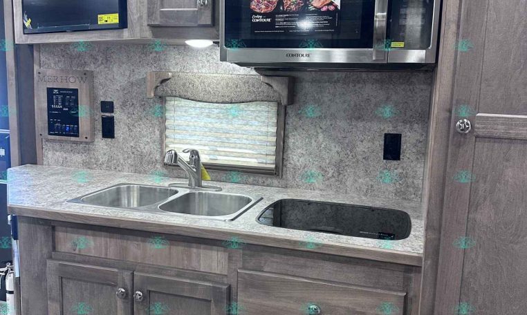 Kitchen area in a trailer: gray wood cabinets, stainless microwave, double sink, and built-in cooktop along a speckled countertop under ceiling lights