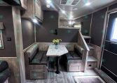 Interior of a modern trailer or RV dining area with a U-shaped booth, marble table, and overhead cabinets in warm wood tones.