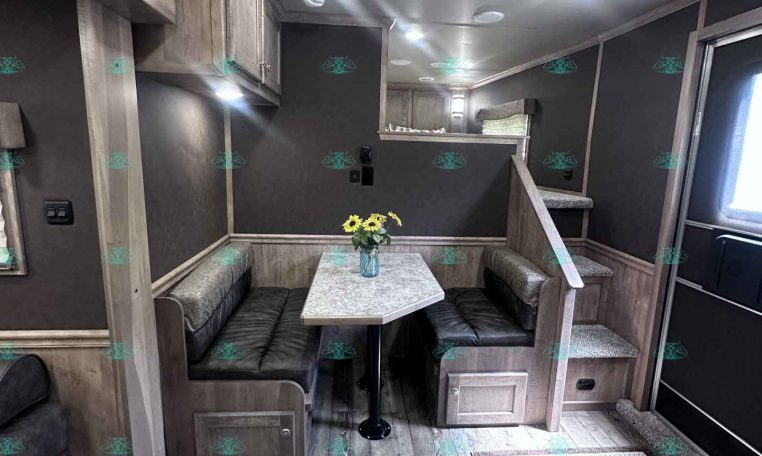 Interior of a modern trailer or RV dining area with a U-shaped booth, marble table, and overhead cabinets in warm wood tones.
