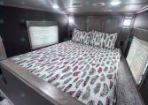 Bedroom inside a trailer: queen bed with colorful feather-pattern quilt, dark walls, and small windows with blinds on each side, ceiling lights above