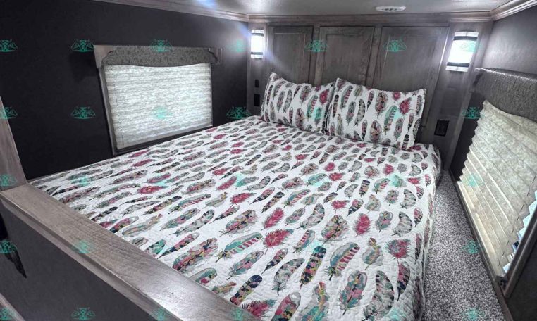 Bedroom inside a trailer: queen bed with colorful feather-pattern quilt, dark walls, and small windows with blinds on each side, ceiling lights above