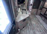 Interior of a modern travel trailer kitchen: wood cabinetry, dual sink, microwave above counter, and a doorway to a small bathroom in the background.