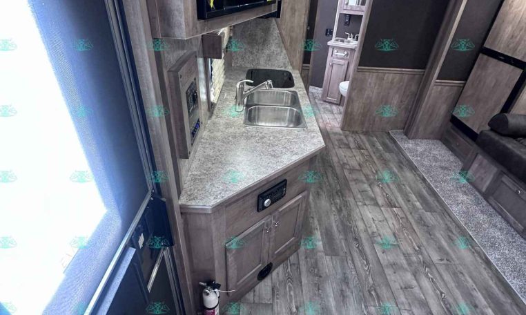Interior of a modern travel trailer kitchen: wood cabinetry, dual sink, microwave above counter, and a doorway to a small bathroom in the background.