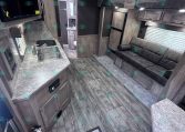 Interior of a modern travel trailer: kitchen on the left with a double sink, microwave, and gray cabinetry; a gray sofa along the right wall; wood-look floors and ceiling lights visible.
