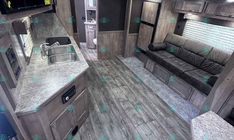Interior of a modern travel trailer: kitchen on the left with a double sink, microwave, and gray cabinetry; a gray sofa along the right wall; wood-look floors and ceiling lights visible.