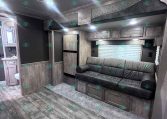Interior of a modern trailer living area with a gray sofa, wood cabinets, and a small kitchen visible in the background.