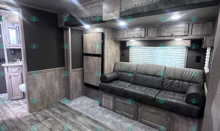 Interior of a modern trailer living area with a gray sofa, wood cabinets, and a small kitchen visible in the background.