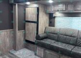 Interior of a modern RV living area with gray wood cabinets, a sofa, and a tall refrigerator beside a pillar and window blinds