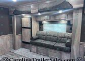 Living area of a travel trailer with gray wood cabinets, a gray leather sofa, and an upper bunk labeled Stampede by Merhow above. A window with blinds is in the background.