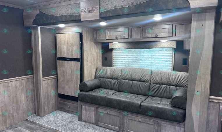 Living area of a travel trailer with gray wood cabinets, a gray leather sofa, and an upper bunk labeled Stampede by Merhow above. A window with blinds is in the background.