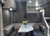 Interior of a camper trailer: U-shaped booth seating around a rectangular table with a vase of sunflowers on top, wood cabinets, and a loft/bed area in the back