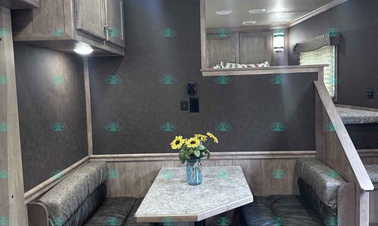 Interior of a camper trailer: U-shaped booth seating around a rectangular table with a vase of sunflowers on top, wood cabinets, and a loft/bed area in the back