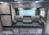 Interior of a travel trailer living area with gray wood cabinets, a gray sofa, and a large window with blinds.
