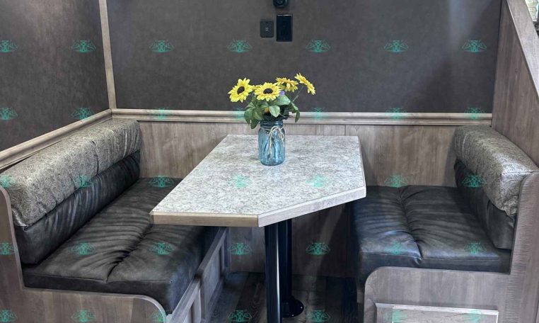 Booth-style dining area in a trailer with gray upholstered benches, a light gray speckled table, and a vase of yellow sunflowers in the center.