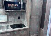 Compact RV kitchen: stainless microwave above a speckled countertop with sink on the left and a black glass induction cooktop, surrounded by gray wood cabinets and a tall pantry.