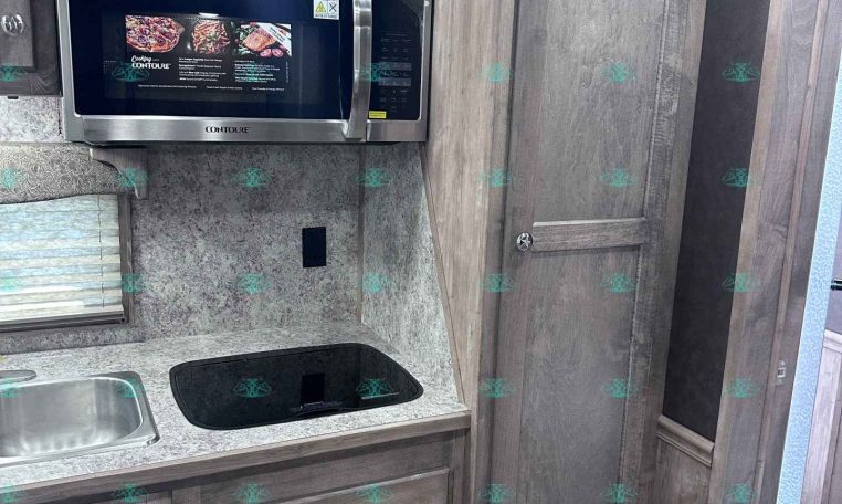 Compact RV kitchen: stainless microwave above a speckled countertop with sink on the left and a black glass induction cooktop, surrounded by gray wood cabinets and a tall pantry.