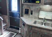 RV interior kitchen: entry door on left, dark wood cabinets, microwave above counter, and double sink with window nearby.