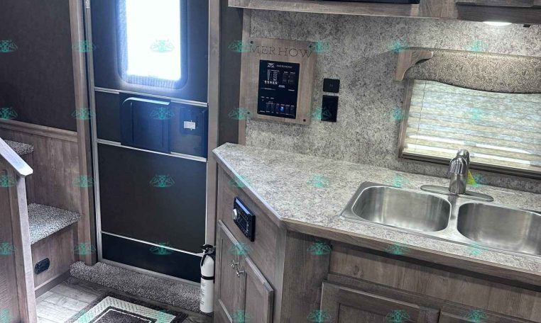 RV interior kitchen: entry door on left, dark wood cabinets, microwave above counter, and double sink with window nearby.