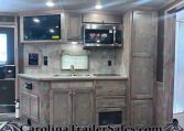 Kitchen interior of a travel trailer with wood cabinets, stainless microwave, sink, and window shade above the counter.